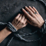 hands bound by a soft velvet rope, symbolizing trust and consensual restraint in BDSM. The background is a muted shade, enhancing the sense of safety and respect intrinsic to BDSM etiquette.